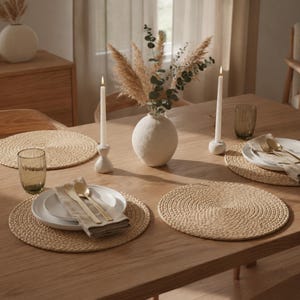 May include: A wooden dining table set with four round woven placemats, white ceramic plates, gold-toned cutlery, beige linen napkins, two white taper candles in ceramic holders, and a central round vase containing dried pampas grass and eucalyptus.