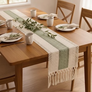 May include: A woven table runner featuring a central sage green stripe flanked by cream-coloured panels with textured patterns and knotted fringe ends. The runner is laid across a wooden table set with white ceramic mugs and plates with napkins.