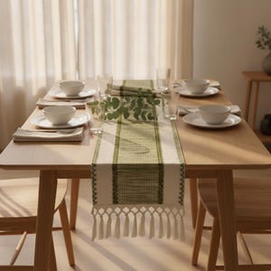 May include: A rectangular table runner featuring a central olive green woven stripe flanked by intricate dark green geometric borders and finished with long, cream-coloured macrame-style fringe tassels at both ends, displayed on a wooden table.