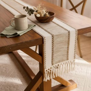 May include: A woven table runner featuring a cream and tan striped pattern with decorative fringe at the ends. The runner is placed on a wooden table alongside a light green cloth napkin, a ceramic mug, and a small wooden bowl with dried flowers.