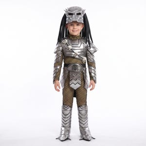May include: A silver and black Predator costume featuring a molded helmet with long black dreadlocks, metallic-colored chest and shoulder armor, a belt with a central emblem, and matching leg guards over a gold-toned mesh bodysuit.