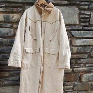 May include: A long, beige canvas coat with a brown corduroy collar and multiple pockets. The coat has a button closure and is lined with a brown fabric.
