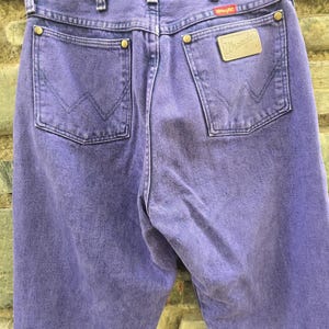 Może przedstawiać: Zbliżenie fioletowych dżins&oacute;w Wrangler. Dżinsy mają dwie tylne kieszenie, sk&oacute;rzaną naszywkę z logo Wrangler i czerwoną metkę. Dżinsy wiszą na wieszaku.
