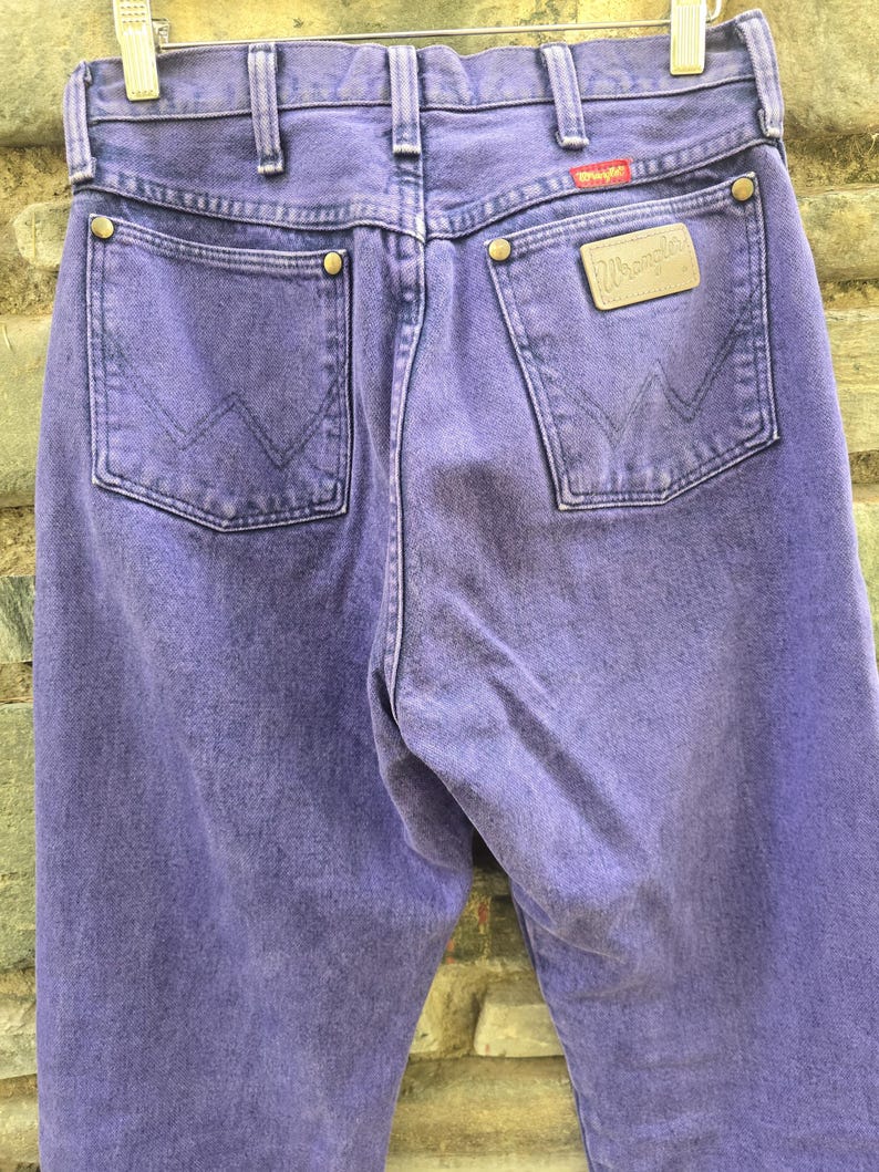 Może przedstawiać: Zbliżenie fioletowych dżins&oacute;w Wrangler. Dżinsy mają dwie tylne kieszenie, sk&oacute;rzaną naszywkę z logo Wrangler i czerwoną metkę. Dżinsy wiszą na wieszaku.