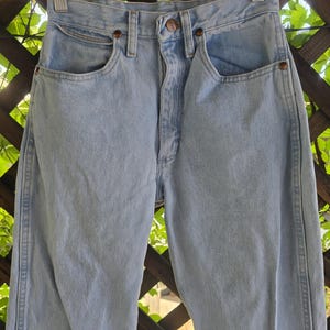 May include: High-waisted light blue denim jeans featuring a classic five-pocket design, a metal button closure at the waist, and copper-toned rivets. The denim has a faded wash finish with visible stitching along the seams and a structured waistband.