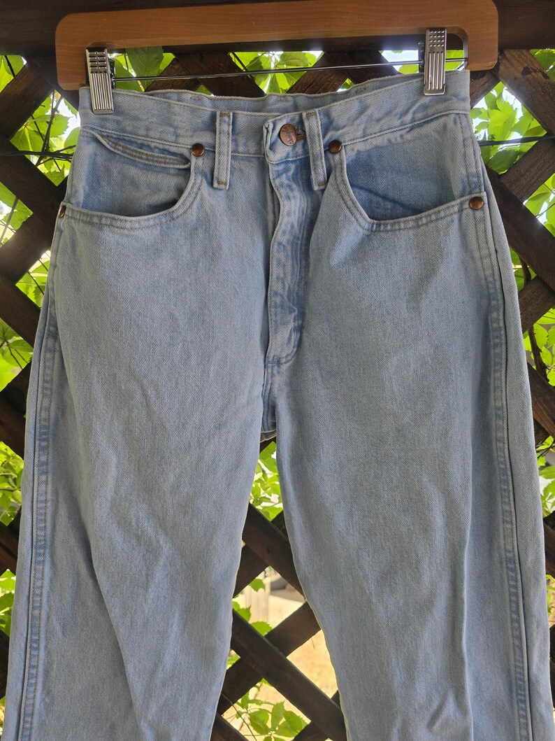 May include: High-waisted light blue denim jeans featuring a classic five-pocket design, a metal button closure at the waist, and copper-toned rivets. The denim has a faded wash finish with visible stitching along the seams and a structured waistband.