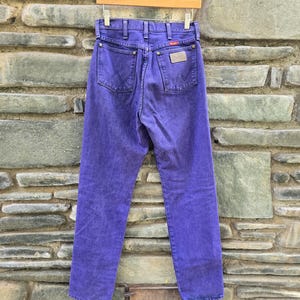 Może przedstawiać: Fioletowe dżinsy z denimu wiszące na drewnianym wieszaku. Dżinsy mają klasyczny pięciokieszeniowy design, metkę Wrangler i wyblakły wygląd. Dżinsy są prezentowane na tle teksturowanej kamiennej ściany.