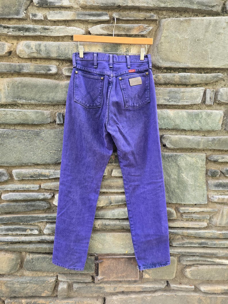 Może przedstawiać: Fioletowe dżinsy z denimu wiszące na drewnianym wieszaku. Dżinsy mają klasyczny pięciokieszeniowy design, metkę Wrangler i wyblakły wygląd. Dżinsy są prezentowane na tle teksturowanej kamiennej ściany.