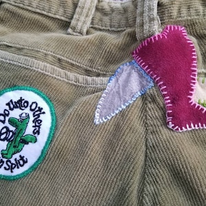 May include: A pair of brown corduroy pants with a green patch that says "Do Unto Others Then Split" and a pink, blue, and white fabric patch sewn on the back.