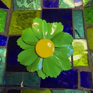 May include: A metal flower ornament featuring layered light green petals with scalloped edges and a smooth, circular yellow center. The flower is positioned against a background of blue and green glass mosaic tiles with dark grout lines.