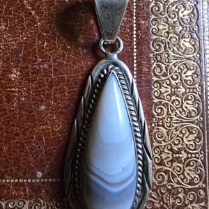 May include: A silver pendant with a teardrop-shaped, light blue and white stone. The stone is encased in a silver setting with a rope-like border and a decorative bail. The pendant is displayed on a brown, patterned surface.