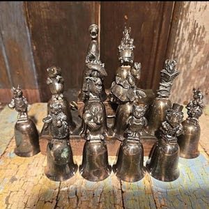 May include: A collection of antique silver-toned bells, each topped with a detailed figurine. The figurines depict various animals and characters, including a bear, owl, and rabbit. The bells are arranged on a wooden surface.