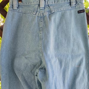 May include: Light blue denim jeans featuring a high-waisted design with a back yoke, belt loops, and a black Wrangler brand tag on the right rear pocket area. The jeans are suspended from a wooden hanger with metal clips at the waistband.