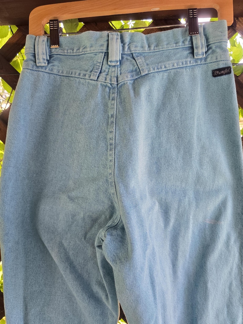 May include: Light blue denim jeans featuring a high-waisted design with a back yoke, belt loops, and a black Wrangler brand tag on the right rear pocket area. The jeans are suspended from a wooden hanger with metal clips at the waistband.