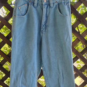 May include: Light blue denim jeans featuring a high-waisted design, a button and zipper fly closure, belt loops, and two front pockets with reinforced metal rivets. The pants have a straight-leg cut and a classic light wash finish throughout.