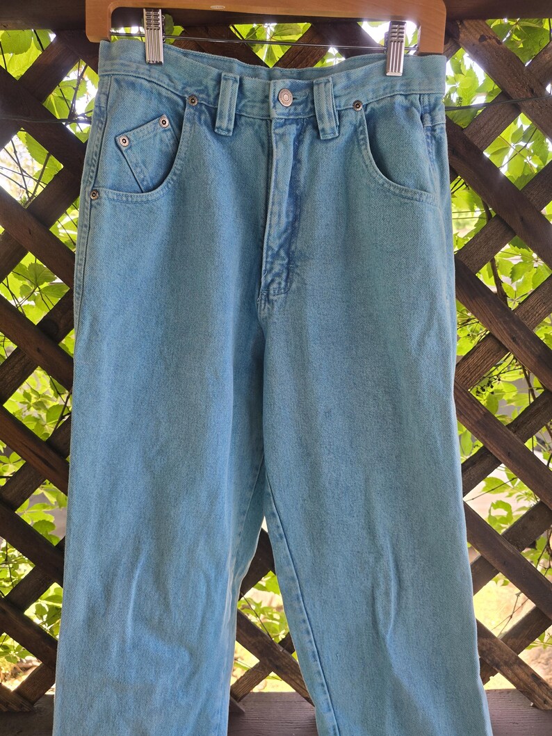 May include: Light blue denim jeans featuring a high-waisted design, a button and zipper fly closure, belt loops, and two front pockets with reinforced metal rivets. The pants have a straight-leg cut and a classic light wash finish throughout.