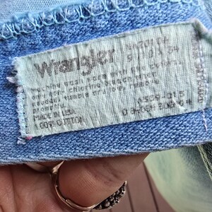 May include: A light blue denim garment featuring a rectangular white fabric tag with the Wrangler logo in black text. The tag includes care instructions, the text Made in USA, and 100% Cotton, stitched onto the interior waistband area.