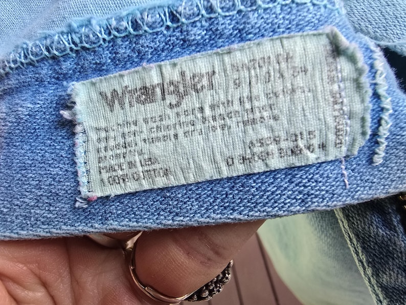 May include: A light blue denim garment featuring a rectangular white fabric tag with the Wrangler logo in black text. The tag includes care instructions, the text Made in USA, and 100% Cotton, stitched onto the interior waistband area.