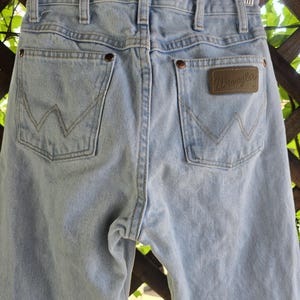 May include: Light blue denim jeans featuring a high-waisted design with belt loops, two back pockets with a signature W-shaped stitch pattern, copper-toned rivets, and a brown leather-like patch on the right pocket with the brand name Wrangler.