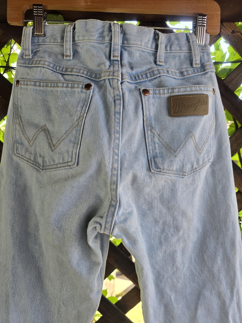 May include: Light blue denim jeans featuring a high-waisted design with belt loops, two back pockets with a signature W-shaped stitch pattern, copper-toned rivets, and a brown leather-like patch on the right pocket with the brand name Wrangler.