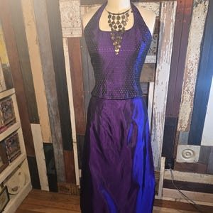 May include: A purple halter top with a black bodice and a matching purple skirt. The top has a square neckline and is adorned with small sequins. The skirt is made of a silky fabric and falls to the floor. The top is paired with a gold coin necklace.