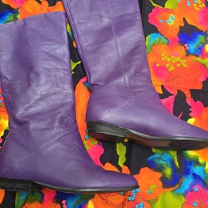 May include: A pair of mid-calf boots crafted from smooth purple leather. These boots feature a rounded toe, a flat black heel, and vertical seam detailing running up the shaft. The leather has a soft, matte finish with visible natural creasing.