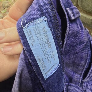 Może przedstawiać: Zbliżenie fioletowych dżins&oacute;w Wrangler. Dżinsy mają niebieską metkę z logo Wrangler i instrukcją prania. Dżinsy są wykonane w 100% z bawełny i mają rozmiar 7x32.