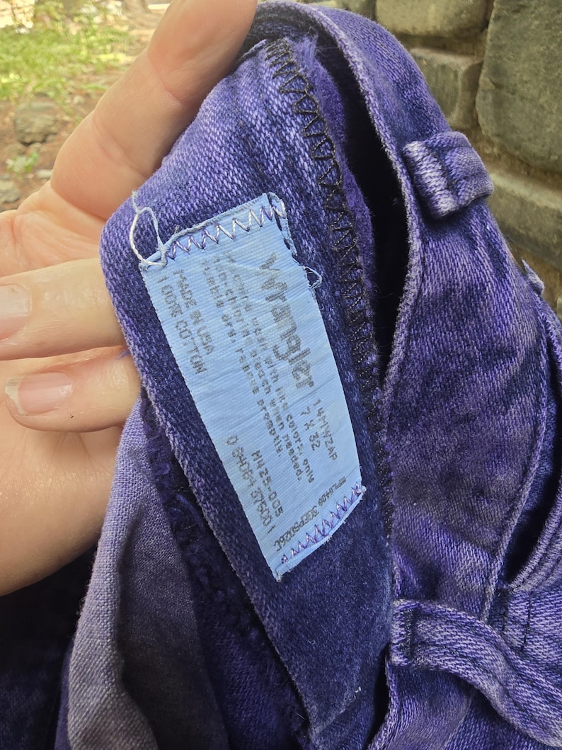 Może przedstawiać: Zbliżenie fioletowych dżins&oacute;w Wrangler. Dżinsy mają niebieską metkę z logo Wrangler i instrukcją prania. Dżinsy są wykonane w 100% z bawełny i mają rozmiar 7x32.