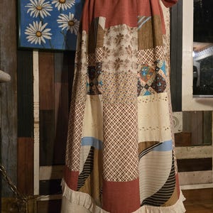 May include: A long, patchwork skirt with a rust-colored waistband and a cream-colored hem with lace trim. The skirt features various fabric patterns in shades of brown, beige, and blue. The skirt is displayed on a mannequin.