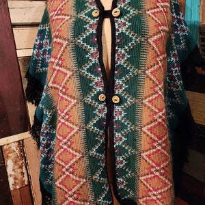 May include: A green, brown, and white patterned poncho with black fringe. The poncho has a black trim and two wooden buttons.