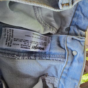 May include: Light blue denim jeans featuring a white interior care label with the Wrangler logo, size 7 x 32, and washing instructions. The jeans have a metal button closure, visible stitching, and a light wash finish on the cotton fabric.