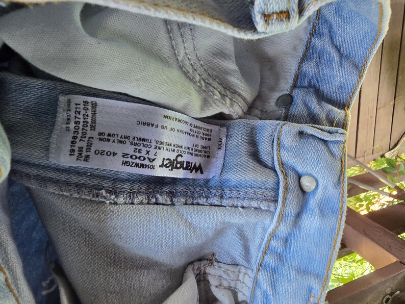 May include: Light blue denim jeans featuring a white interior care label with the Wrangler logo, size 7 x 32, and washing instructions. The jeans have a metal button closure, visible stitching, and a light wash finish on the cotton fabric.