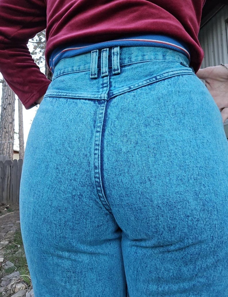 May include: A pair of high-waisted blue jeans with a light blue acid wash finish. The jeans have a back pocket detail with two vertical seams.
