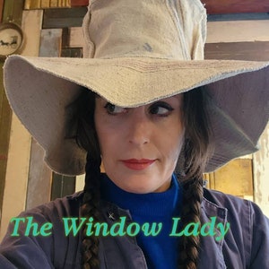May include: A wide-brimmed, beige hat with a frayed edge. The hat is worn with a blue turtleneck and a dark blue jacket. The text "The Window Lady" is displayed in green script at the bottom of the image.
