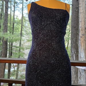 May include: A one-shoulder, navy blue, sparkly dress with a swirling pattern. The dress is fitted and has a long, flowing skirt.