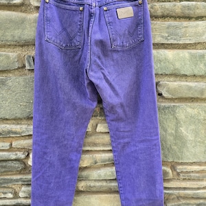 Może przedstawiać: Para fioletowych dżins&oacute;w Wrangler, widocznych z tyłu, wiszących na wieszaku. Dżinsy mają dwie tylne kieszenie, sk&oacute;rzaną naszywkę i logo Wrangler. Denim jest jednolity i żywy fioletowy.