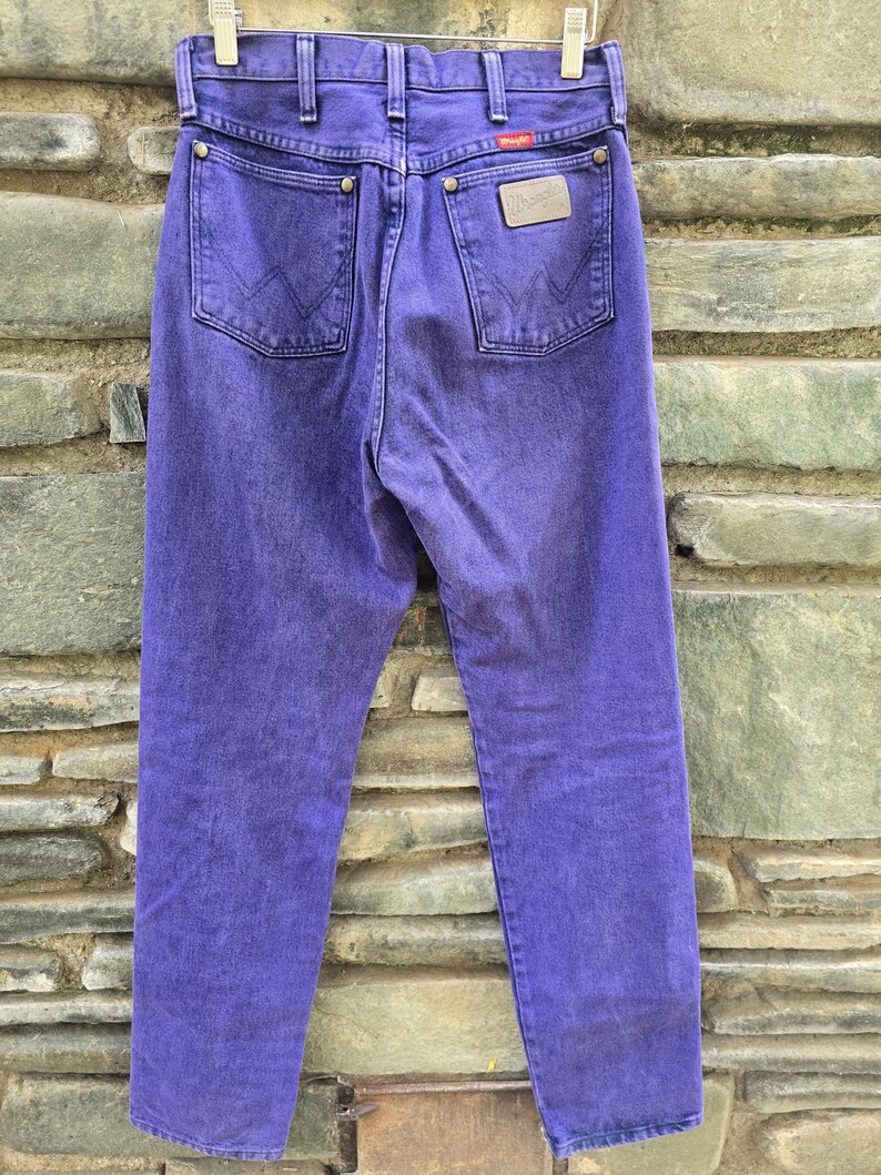 Może przedstawiać: Para fioletowych dżins&oacute;w Wrangler, widocznych z tyłu, wiszących na wieszaku. Dżinsy mają dwie tylne kieszenie, sk&oacute;rzaną naszywkę i logo Wrangler. Denim jest jednolity i żywy fioletowy.