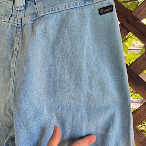 May include: Light blue denim jeans featuring a high-waisted design with belt loops and a black Wrangler brand tag on the back pocket area. The fabric has a light wash finish and shows a small, faint, light-colored mark on the lower right side.