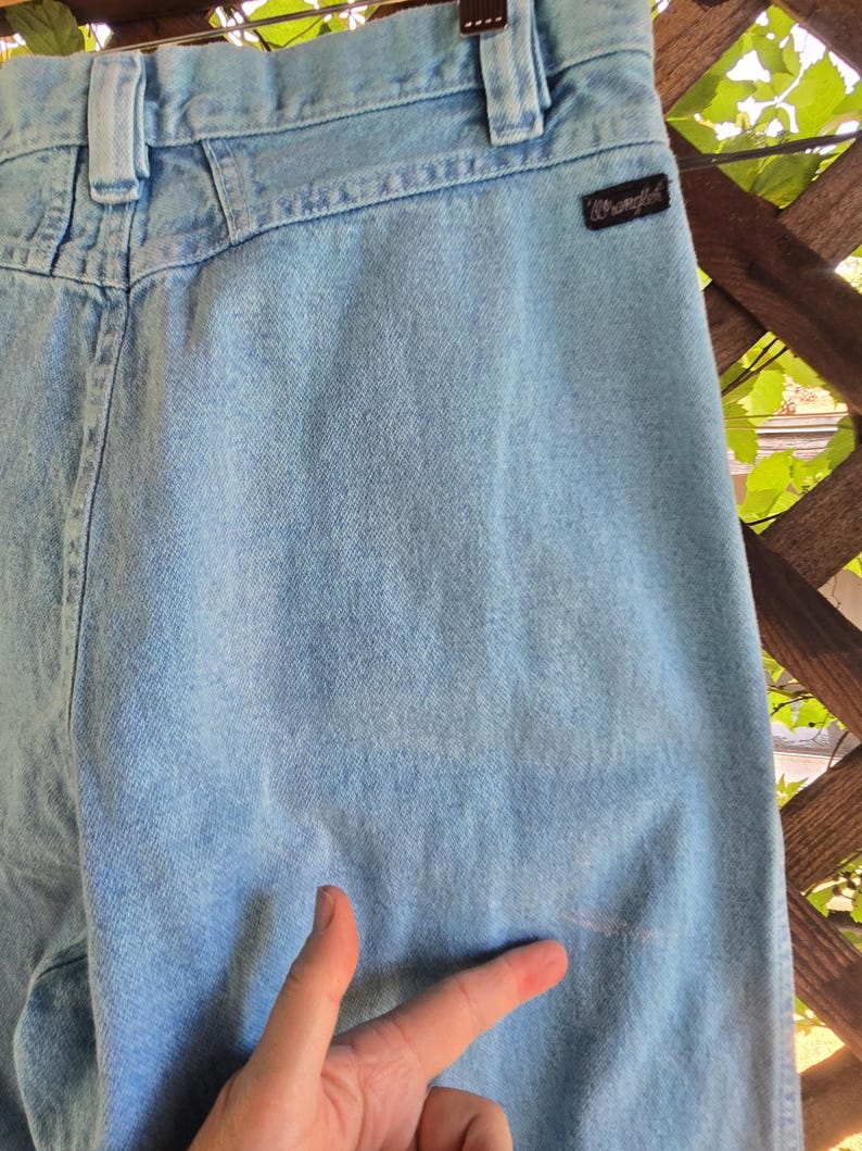 May include: Light blue denim jeans featuring a high-waisted design with belt loops and a black Wrangler brand tag on the back pocket area. The fabric has a light wash finish and shows a small, faint, light-colored mark on the lower right side.