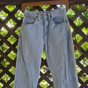 May include: Light blue denim jeans featuring a high-waisted design, a button fly closure, two front pockets, and a small coin pocket. The pants have a straight-leg cut with visible stitching along the side seams and copper-toned metal rivets.