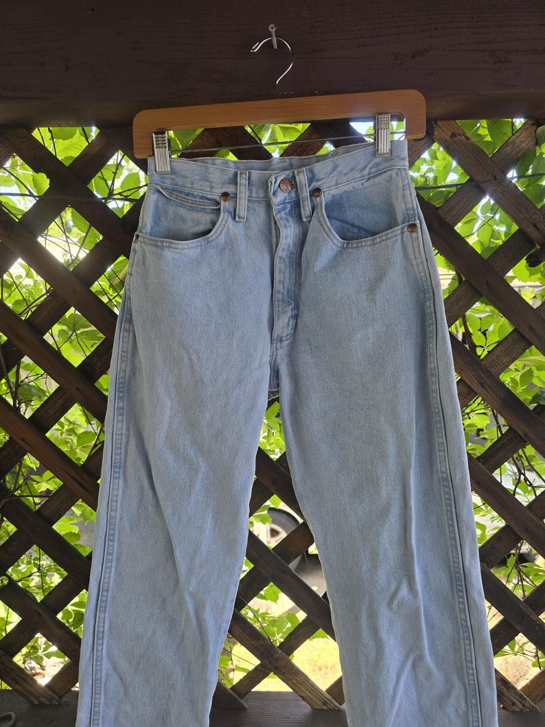 May include: Light blue denim jeans featuring a high-waisted design, a button fly closure, two front pockets, and a small coin pocket. The pants have a straight-leg cut with visible stitching along the side seams and copper-toned metal rivets.