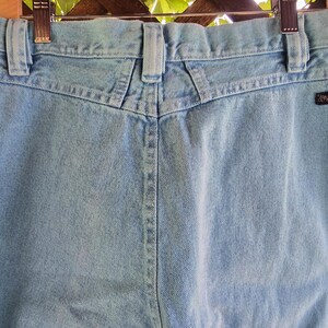 May include: Light blue denim jeans featuring a high-waisted design with a V-shaped back yoke, belt loops, and a black Wrangler brand tag on the right rear pocket area. The jeans are constructed from light-wash cotton denim fabric with visible stitching.