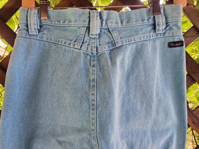 May include: Light blue denim jeans featuring a high-waisted design with a V-shaped back yoke, belt loops, and a black Wrangler brand tag on the right rear pocket area. The jeans are constructed from light-wash cotton denim fabric with visible stitching.