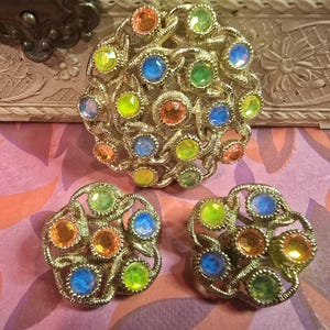 May include: A set of three gold-tone brooches featuring colourful rhinestones. The largest brooch is circular with a floral pattern. The two smaller brooches are round and have a similar floral design.