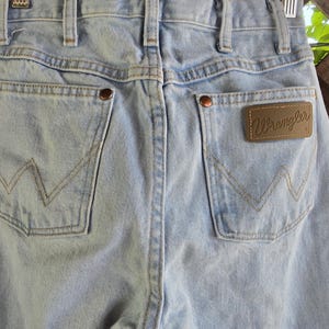 May include: Light blue denim jeans featuring a high-waisted design with belt loops, two back pockets with copper-colored rivets, and a brown leather patch on the right pocket with the word Wrangler embossed in a cursive script font.