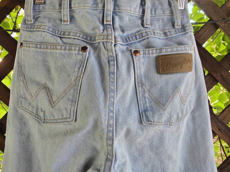 May include: Light blue denim jeans featuring a high-waisted design with belt loops, two back pockets with copper-colored rivets, and a brown leather patch on the right pocket with the word Wrangler embossed in a cursive script font.