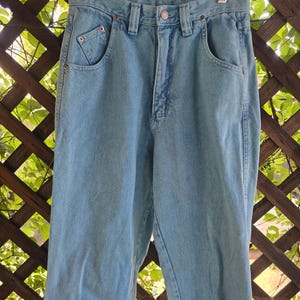 May include: High-waisted light blue denim jeans featuring a classic five-pocket design, a metal button closure, a zippered fly, and belt loops. The jeans have a straight-leg cut with visible copper-toned rivets at the pocket corners.