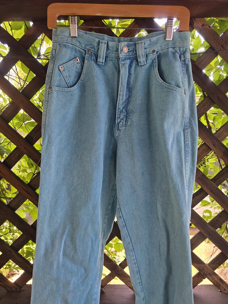 May include: High-waisted light blue denim jeans featuring a classic five-pocket design, a metal button closure, a zippered fly, and belt loops. The jeans have a straight-leg cut with visible copper-toned rivets at the pocket corners.