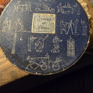 May include: A round, dark blue box with a vintage design featuring white line drawings of Parisian scenes. The box has the words "Evening in Paris" on a silver label. The box is decorated with lace and ribbon.