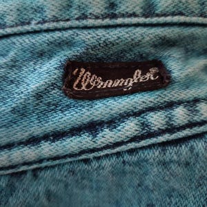 May include: Close-up of a blue denim fabric with a black leather patch that says "Wrangler" in white lettering.